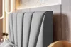 A close-up view of a grey upholstered headboard with vertical channel tufting, set against a wooden wall. Náhled obrázku produktu