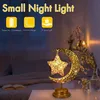 A decorative night light shaped like a crescent moon with a star inside, placed on a table in a bedroom setting. Produktbild-Vorschau