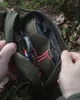 Hands holding open a green hip pack containing a compass and other items, set against a forest floor background. Produktbild-Vorschau