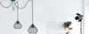 A minimalist studio setting with a white wall background, featuring two black spider pendant lights, a grid panel, a potted plant with yellow fruit, and a studio spotlight. Produktbild-Vorschau