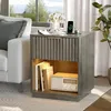 A nightstand with a drawer and an open shelf, placed next to a sofa and near a window. A phone is charging on top, and books and a radio are on the shelf. Anteprima dell’immagine prodotto