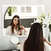 A woman is brushing her hair in front of a lighted mirror. Produktbild-Vorschau
