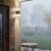An outdoor wall lamp illuminates a porch area with a table and chairs, set against a misty garden backdrop with falling leaves. Produktbild-Vorschau