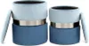 Two cylindrical ottomans with lids, one slightly smaller than the other, are shown against a white background. The ottomans have a two-tone design with a light blue top section and a darker blue bottom section, separated by a silver band. Produktbild-Vorschau