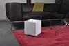 A white cube-shaped humidifier sits on a red rug in front of a dark brown leather sofa. A yellow throw blanket is draped over the sofa. A black coffee table with a silver base is partially visible to the right. Produktbild-Vorschau