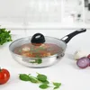 A stainless steel frying pan with a glass lid, filled with food, sits on a white countertop next to some fresh ingredients like a tomato, spinach, garlic, and red onion. Produktbild-Vorschau