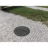 A black manhole cover is placed on a cobblestone path next to a patch of grass. Produktbild-Vorschau