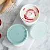 Two ice cream containers with lids, one filled with ice cream and the other empty, sit on a marble surface next to waffle cones and raspberries. Produktbild-Vorschau