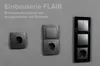 Several electrical outlets and light switches mounted on a textured gray wall, with accompanying text indicating 'Einbauserie FLAIR' and 'Beispielanwendung von Komponenten mit Rahmen'. Produktbild-Vorschau