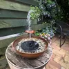 A solar-powered water fountain is set up in a garden, with water spraying into a bowl filled with pebbles and the fountain mechanism. Flowers and greenery surround the fountain, and a chair is visible in the background. Produktbild-Vorschau