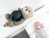 A plush otter toy with a dark blue sphere on its belly lies on a white surface next to a baby in a crib. Produktbild-Vorschau