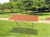 A wooden table with a metal frame is set up on a grassy lawn in a park-like setting with trees in the background. Produktbild-Vorschau