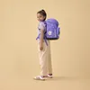 A young girl wearing a purple backpack stands against a plain light yellow background. Produktbild-Vorschau