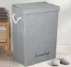 A grey laundry basket with a lid and rope handles sits on a tiled floor next to a wooden shelf with folded towels and a woven basket. Podgląd zdjęcia produktu