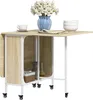 A drop-leaf dining table with storage shelves and wheels, decorated with a vase of flowers, a teacup, and stacked bowls. Produktbild-Vorschau