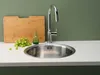 A kitchen sink with a faucet, cutting board, grapes, and a small green bottle on a white countertop against a green wall. Produktbild-Vorschau
