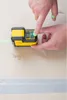 A person's hands are using a yellow and black laser level on a light-colored surface with a strip of tape below. Produktbild-Vorschau