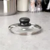 A glass pot lid with a black handle and a stainless steel rim rests on a speckled gray countertop. Náhled obrázku produktu