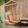 A colorful striped hanging chair is suspended in front of a rustic wooden wall, with greenery and a small stool nearby. Produktbild-Vorschau