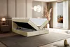 A bedroom scene featuring a beige velvet boxspring bed with its storage compartment open, revealing the mattress and topper. The room has elegant wall paneling, a bedside table with a lamp, and a large window with curtains. Produktbild-Vorschau