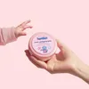 A baby's hand reaches for a pink jar of cream held by an adult hand. Anteprima dell’immagine prodotto