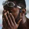 A close-up of a swimmer wearing goggles and bone conduction headphones, covering their face with their hands. Produktbild-Vorschau