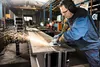 A worker is using an angle grinder to cut a metal beam in a factory setting. Produktbild-Vorschau