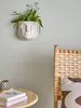 A wall-mounted planter shaped like a face holds a green plant. A wooden chair with a patterned cushion is visible to the right, and a small table with books and a cup is in the foreground. Produktbild-Vorschau