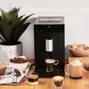 A coffee machine is shown on a wooden counter with coffee beans, sugar cubes, cookies, and cups of coffee. Produktbild-Vorschau