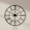 A black metal wall clock with Roman numerals is mounted on a light-colored wall, casting a shadow. Voir les images du produit