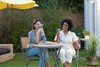 Two women are sitting at a table in a backyard, drinking from portable blenders. Podgląd zdjęcia produktu