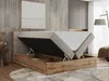 A boxspring bed with its storage compartment open, showcasing the mattress and the wooden frame. The bed is in a bedroom setting with a textured wall and a floor rug. Produktbild-Vorschau