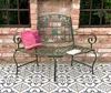 A green metal garden bench with a decorative pillow and a book, placed on a patterned tiled patio in front of a brick wall. Anteprima dell’immagine prodotto