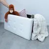 A hand is placing a stuffed monkey on top of a white storage bench. A pink pillow and a fluffy white blanket are also inside the bench. Podgląd zdjęcia produktu