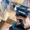 A hand is using a Makita impact driver to screw a metal bracket into wooden beams. Podgląd zdjęcia produktu