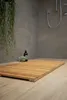 A wooden bath mat is placed on a tiled floor in a modern bathroom, with a showerhead and a plant visible in the background. Produktbild-Vorschau
