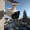 A solar-powered security camera mounted on a textured wall, with a blurred background of houses and a street. Produktbild-Vorschau