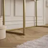 A close-up view of the base of a gold-framed table with a white wall and wooden floor in the background. Produktbild-Vorschau