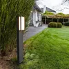 A modern outdoor lamp illuminates a garden path next to lush greenery and a house in the background. Anteprima dell’immagine prodotto