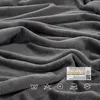 A close-up shot of a gray, soft, textured fabric, likely a blanket or bedding, with folds and creases. A label with certifications and symbols is visible in the lower right corner. Produktbild-Vorschau