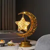 A decorative lamp shaped like a crescent moon with a star hanging from it, placed on a table with cupcakes. Produktbild-Vorschau