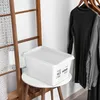 A white storage box with a lid sits on a small round table next to a wooden ladder with a lace-like fabric draped over it. Clothes hang on hangers in the background. Produktbild-Vorschau