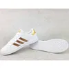 A pair of white Adidas sneakers with gold stripes and accents, displayed on a light gray textured surface. Produktbild-Vorschau