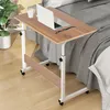 A portable, height-adjustable wooden table with wheels is placed next to a bed, with a laptop and a mug on it. Produktbild-Vorschau