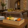 A cheese platter with a wooden base and a clear lid, set on a table with other food items and drinks in the background. Náhľad obrázku produktu