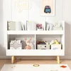 A white children's bookshelf with wooden legs, filled with books and toys, set against a white paneled wall. Náhľad obrázku produktu