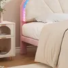 A close-up view of a pink upholstered bed with a beige duvet and a white pillow. The headboard has a decorative fan shape and is illuminated by colorful LED lights. A small white nightstand with glass doors is visible to the left. Náhled obrázku produktu