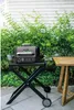 A grill stand with a grill on it, placed on a patio next to a wooden table. Náhľad obrázku produktu
