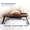 A foldable laptop table is set up on a bed, with a piece of bread on a plate on the table. Blue arrows indicate movement or space around the table. Produktbild-Vorschau
