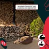 A woven basket filled with firewood is placed on a paved surface in front of a large stack of firewood. There are also green bushes in the background. Voir les images du produit